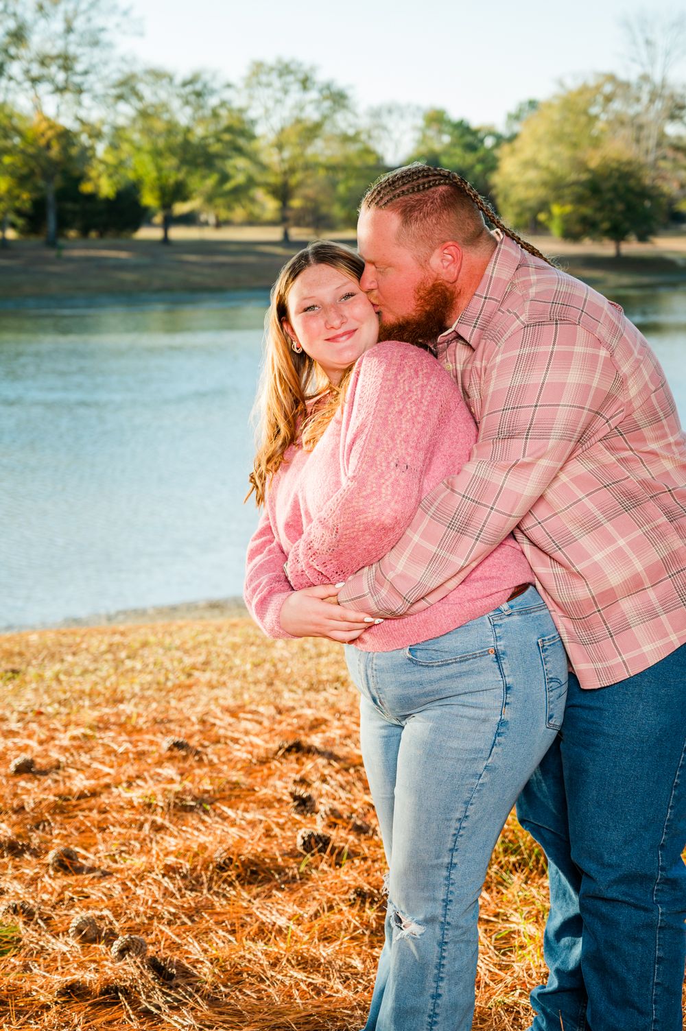 A couple embraces near a lake, the man kissing her cheek, both wearing pink and jeans in a sunny outdoor setting.