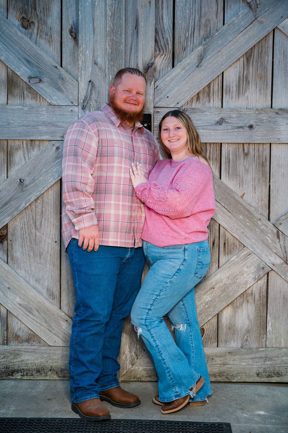 Couple in pink shirts and jeans posing against rustic wooden barn doors, wearing cowboy boots.