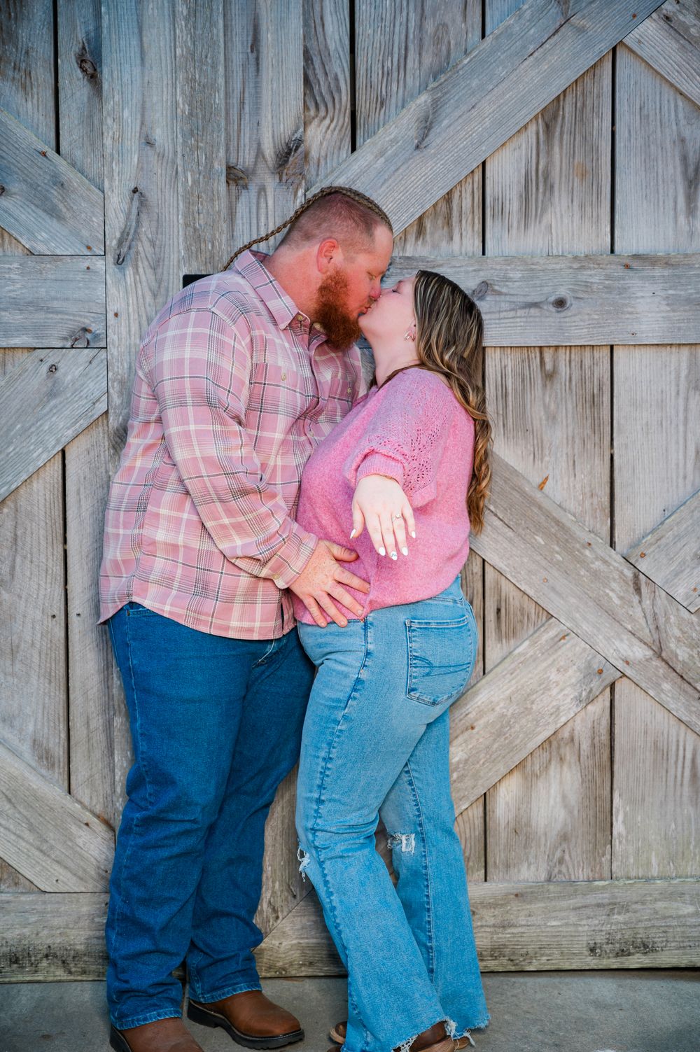 Couple sharing a romantic kiss against a rustic wooden barn door, wearing pink tops and blue jeans.
