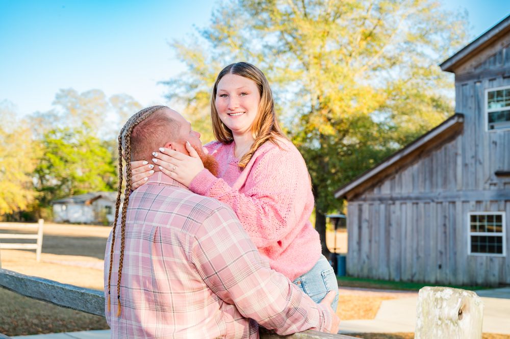 A smiling girl in a pink hoodie being lifted by a man in a plaid shirt outdoors near a rustic barn.