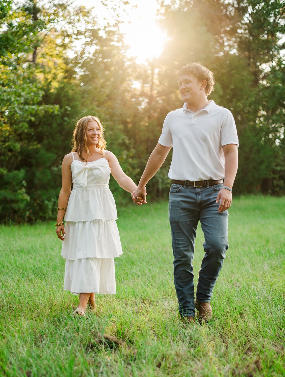 A couple strolls through a green field during golden hour while holding hands.
