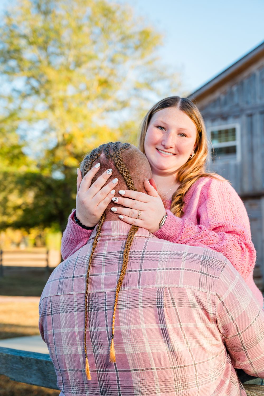 Two women embracing warmly outdoors near a rustic barn surrounded by autumn trees in golden sunlight.
