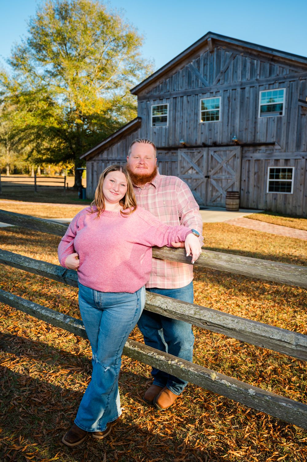 Couple posing by a rustic wooden fence with a gray barn in the background on a sunny fall day.