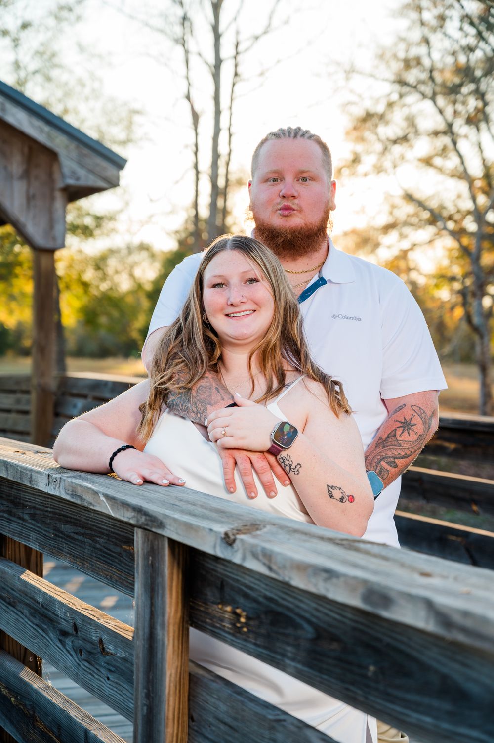 Couple posing on wooden bridge at sunset, woman in white dress leaning on railing, man with tattoos standing behind her.