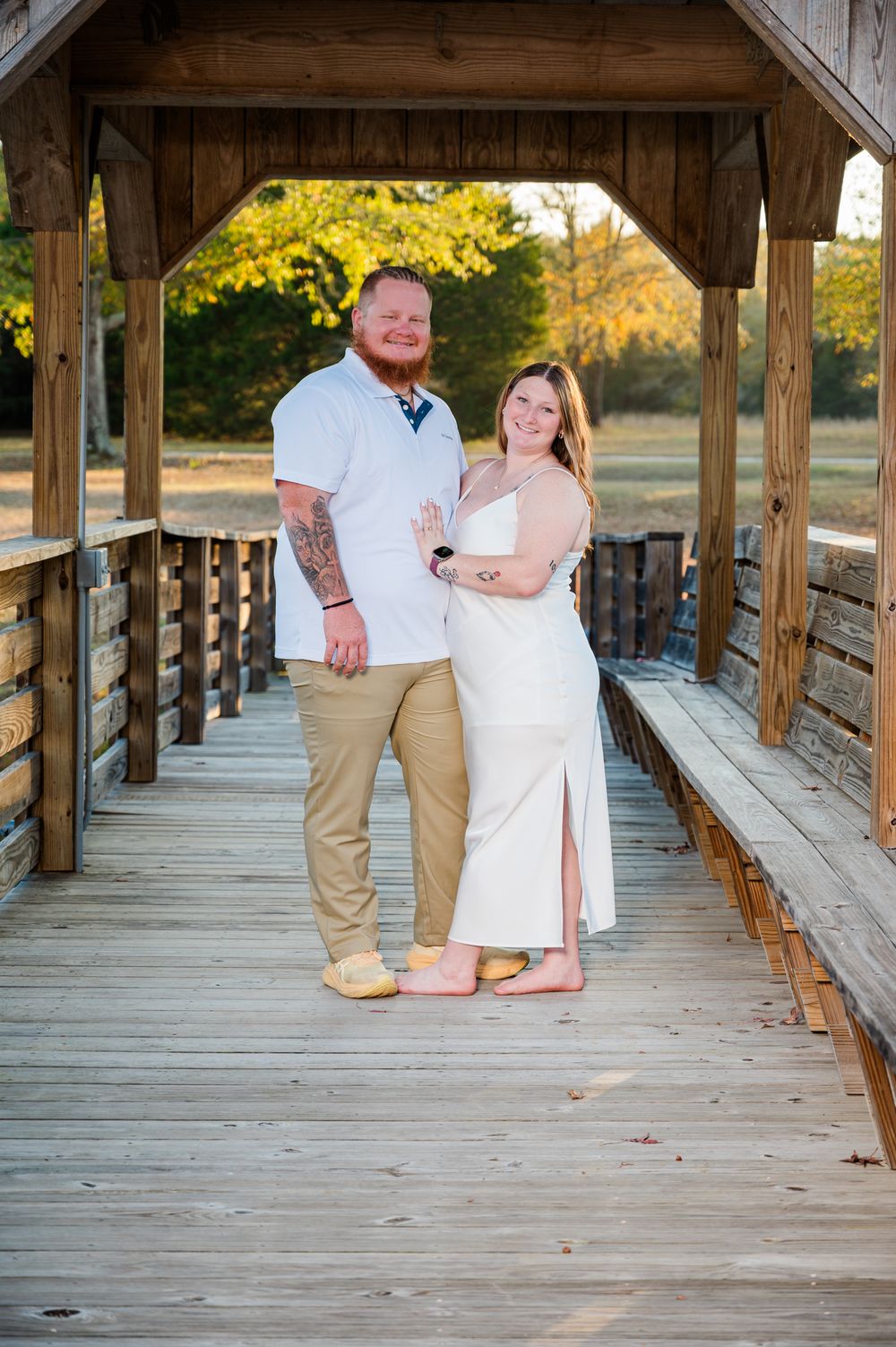 Couple smiling on a wooden covered bridge, man in white shirt and khakis, woman in white dress, autumn trees background.