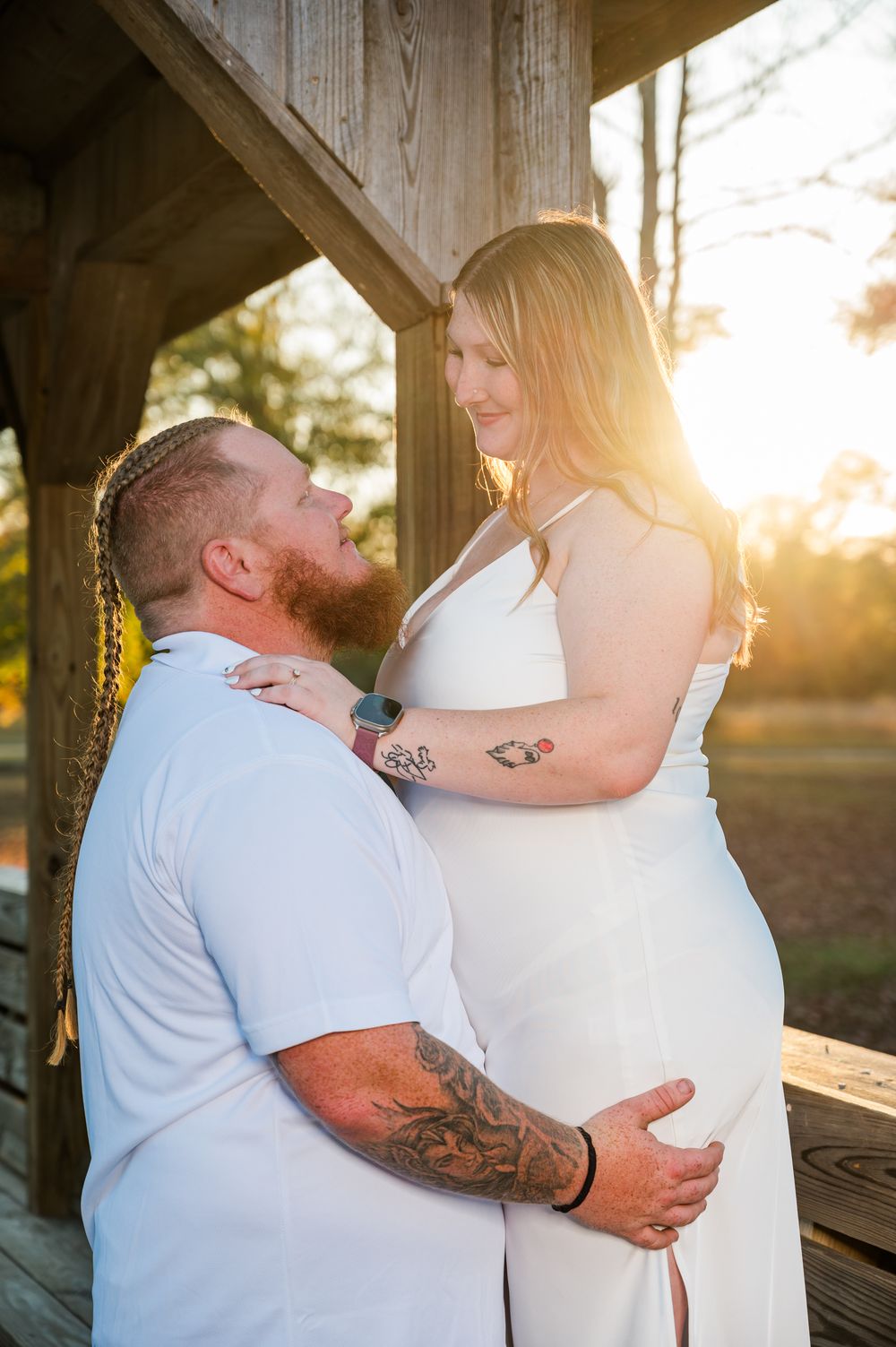Pregnant woman in white dress embraces tattooed man in golden sunset light during maternity photo session.