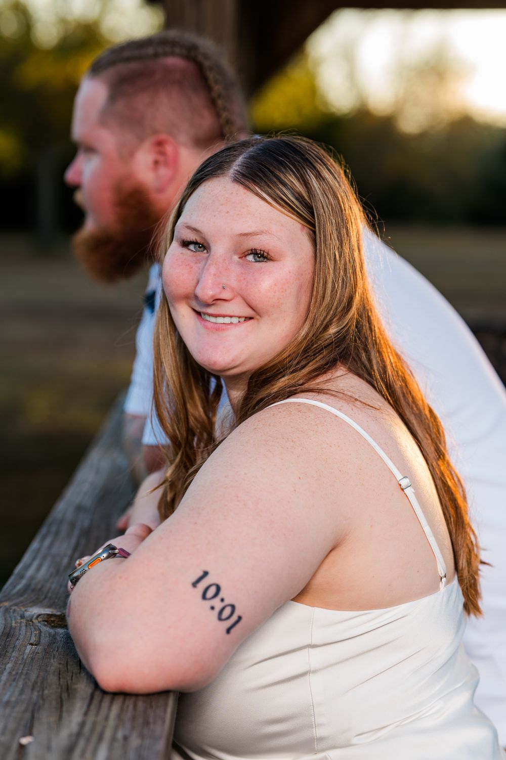 Smiling woman with freckles and a 10:01 tattoo on her arm leans on a wooden railing outdoors at golden hour.