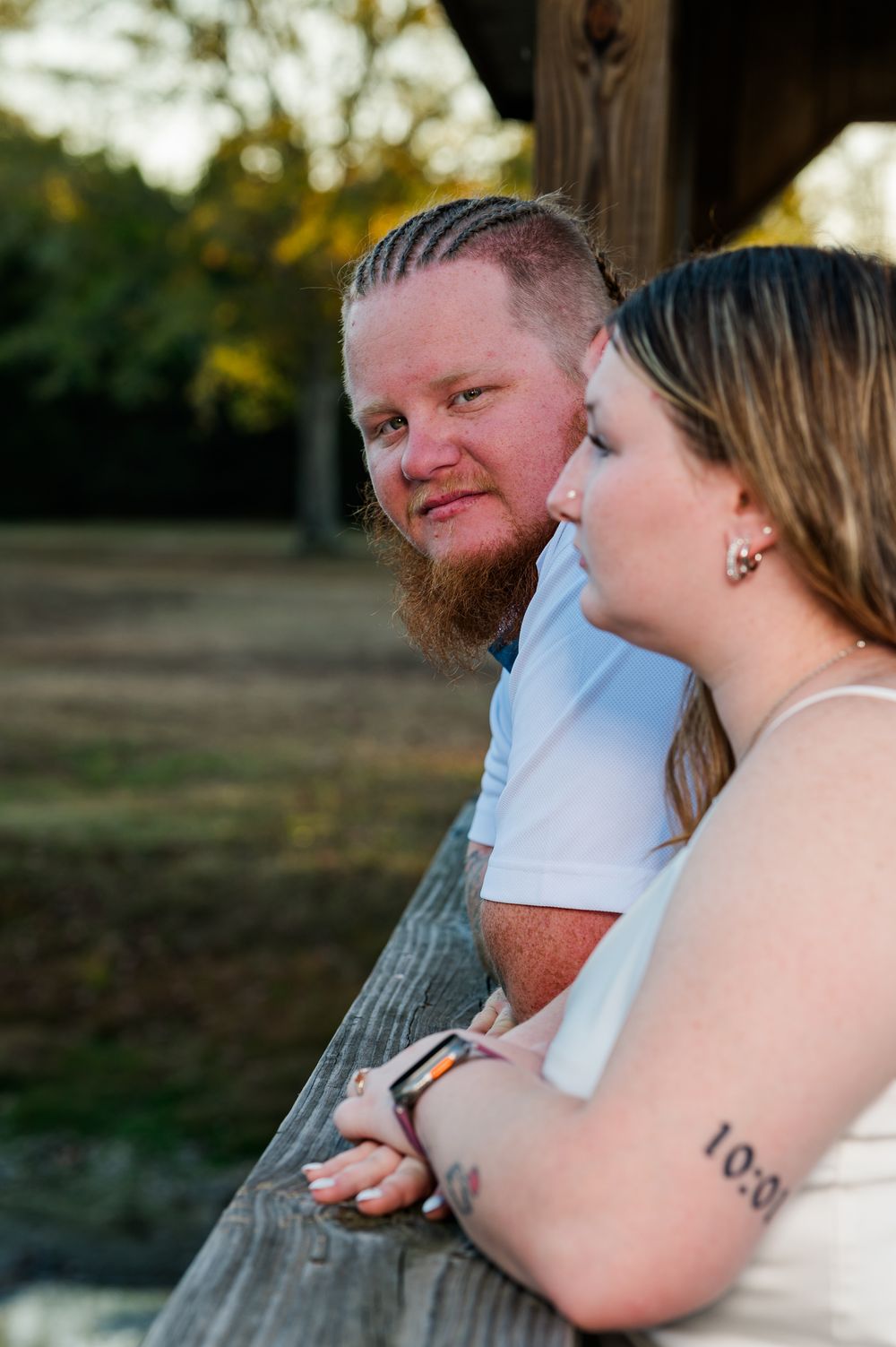 Tattooed couple leans on wooden railing outdoors during golden hour maternity photoshoot.