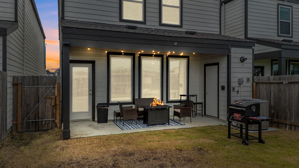 Cozy backyard patio at dusk with string lights, outdoor seating, fire pit, dining table, and a grill on green lawn.