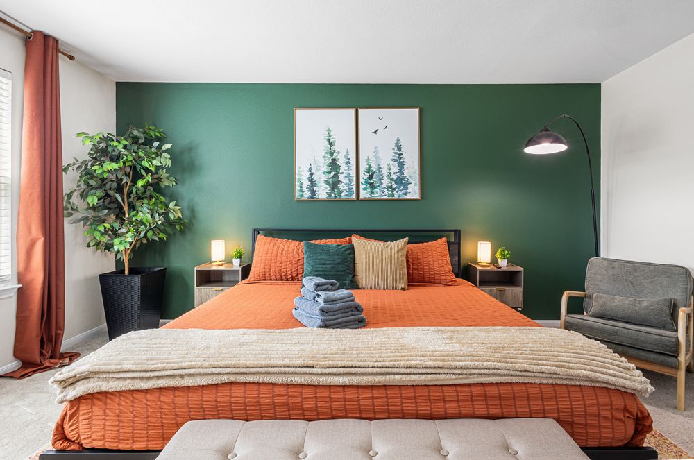 Modern bedroom with green accent wall, orange bedding, framed tree art, potted plant, and cozy armchair.