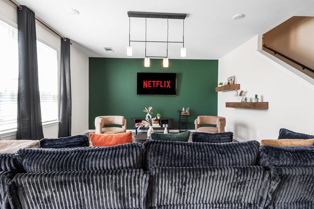 Modern living room with dark sofa, green accent wall, Netflix TV, pendant lighting, and floating shelves.