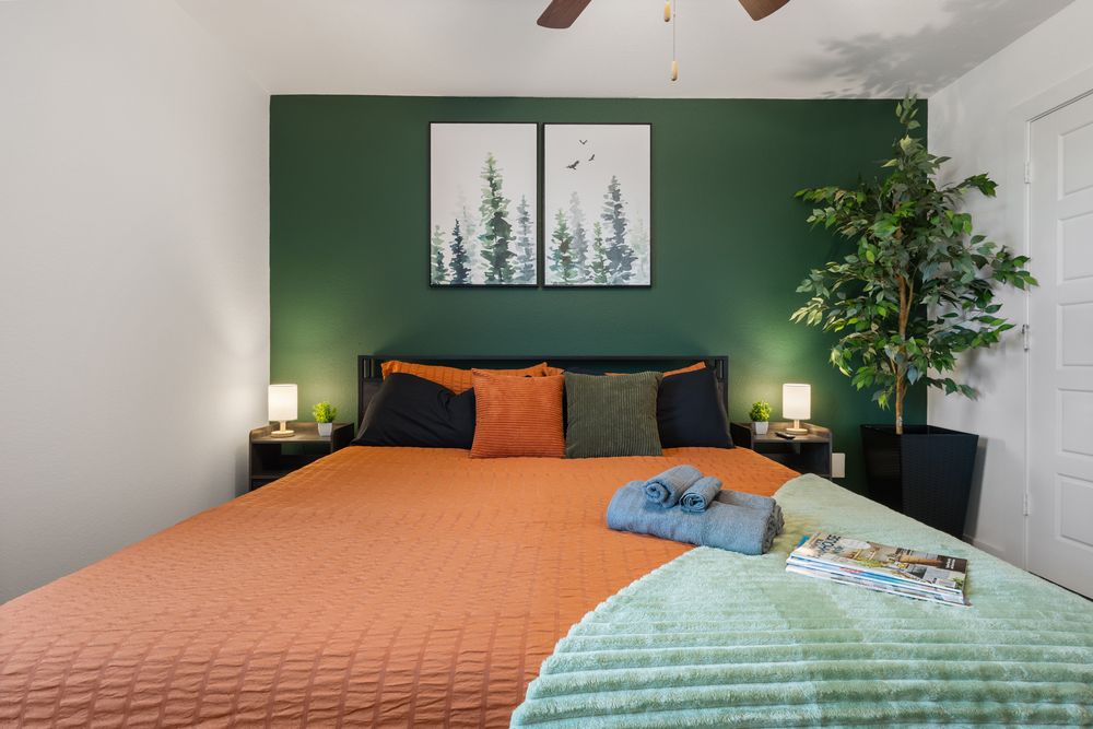 Cozy bedroom with green accent wall, orange bedding, decorative pillows, nightstands with lamps, and a large tree painting above the bed.