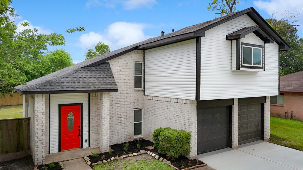 Two-story brick home with red front door, white siding, dark roof, and two-car garage surrounded by lush greenery.