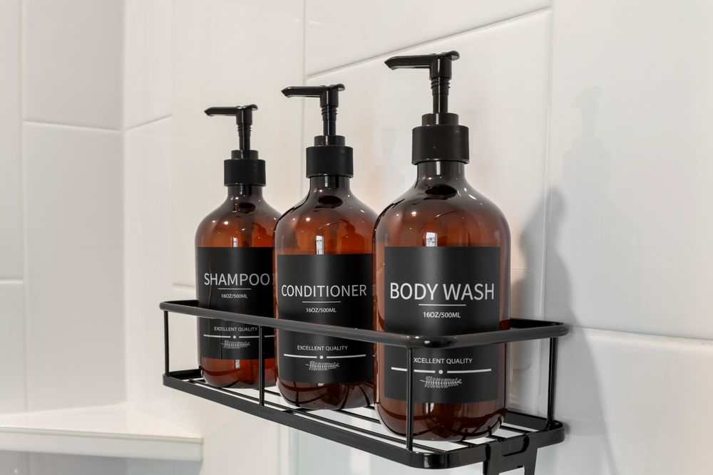 Three amber glass pump bottles labeled Shampoo, Conditioner, and Body Wash on a black metal shelf.
