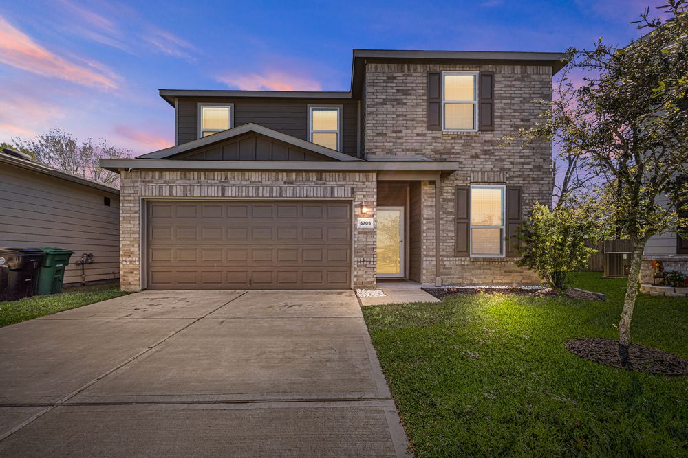 Two-story brick home with a two-car garage, stone accents, and lush landscaping illuminated at dusk.