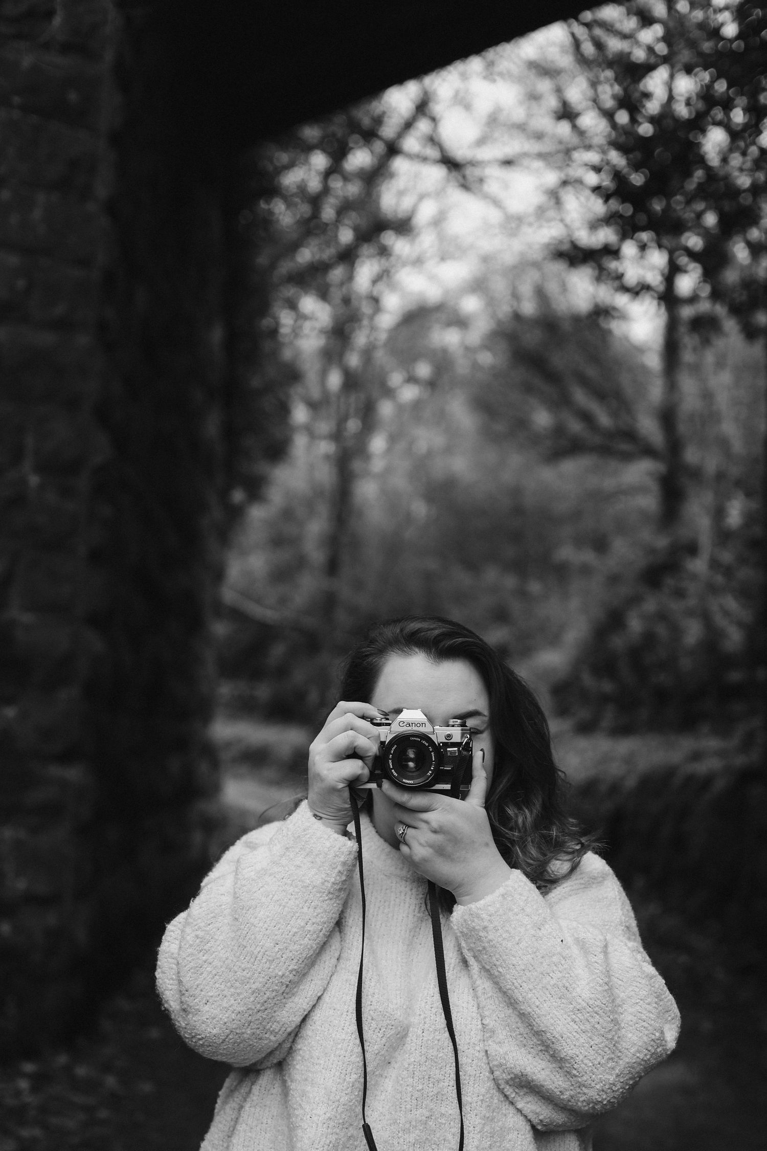 A black and white photo showing someone holding a vintage camera in a wooded setting.