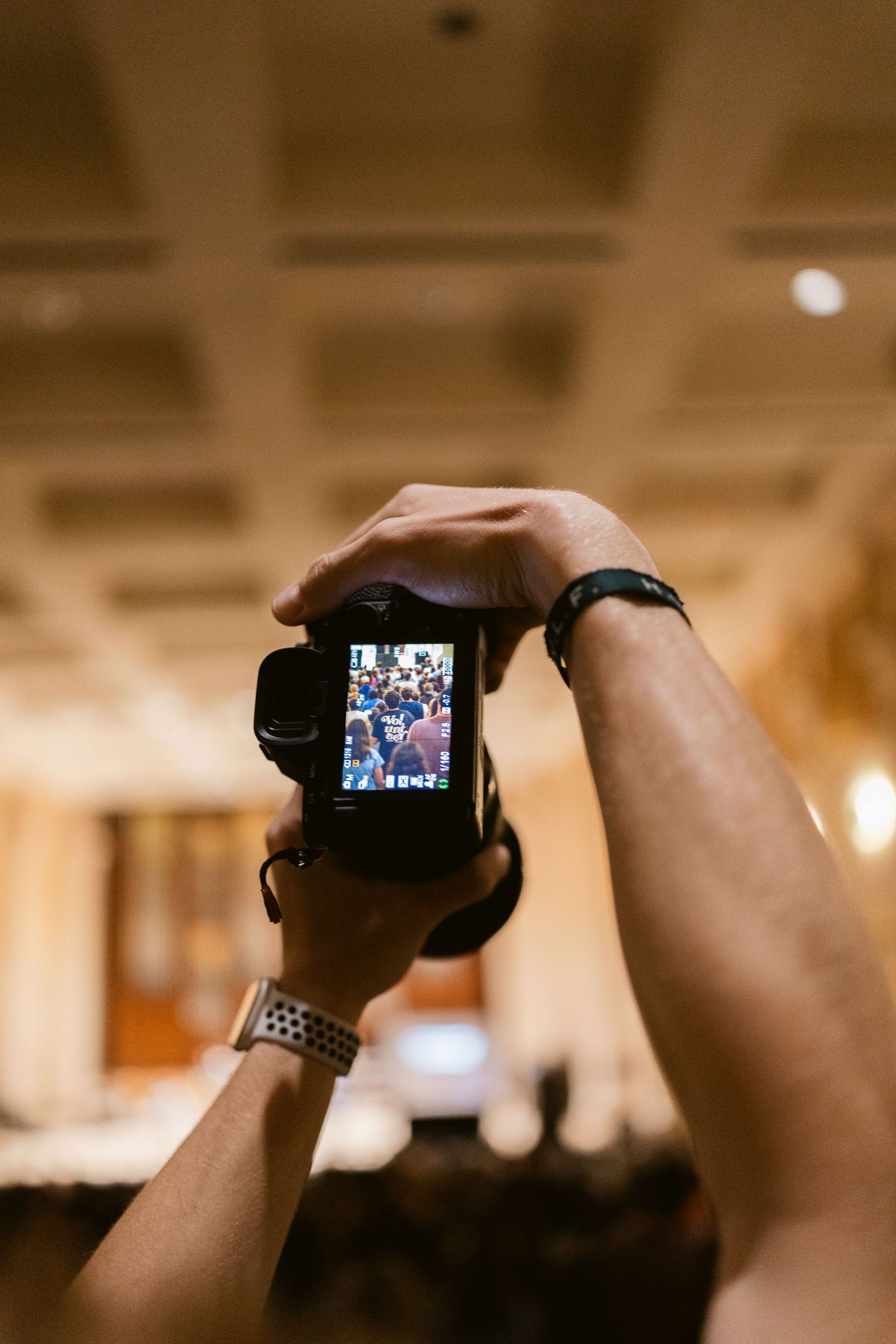 Person holds DSLR camera overhead to photograph a crowded conference or event in a large hall.