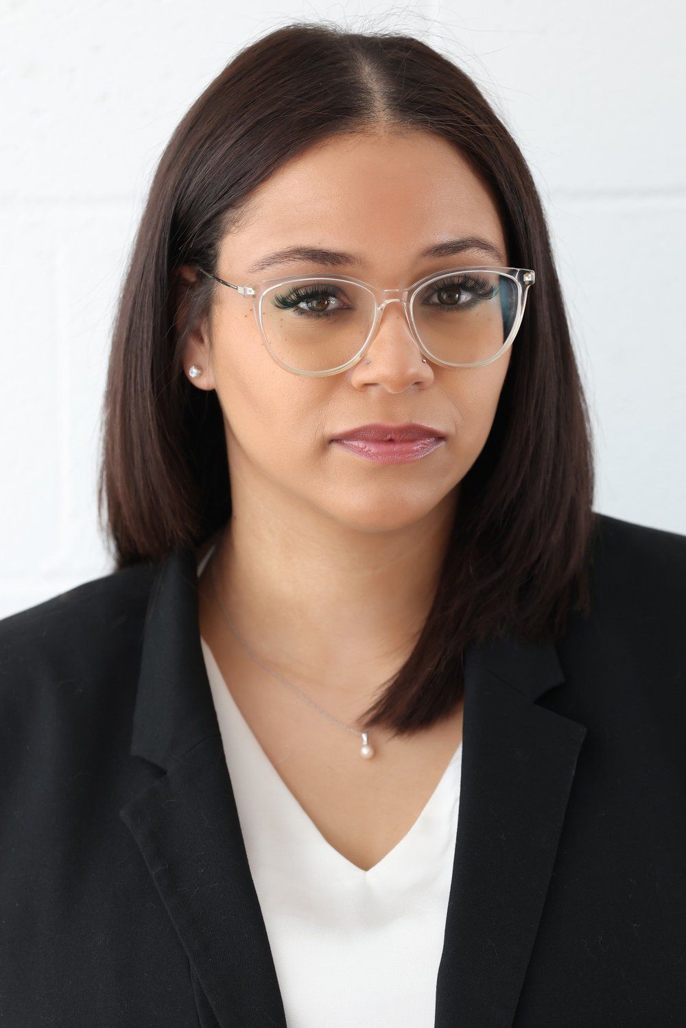 Professional headshot of someone in a black blazer, white blouse and clear glasses against white background.