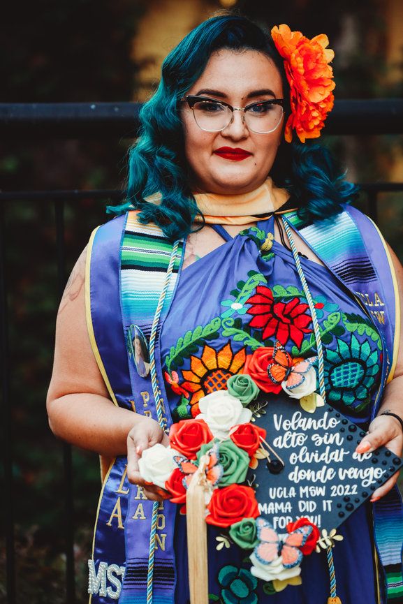 A graduate in vibrant attire wearing glasses and teal hair with an orange flower and colorful graduation decorations.