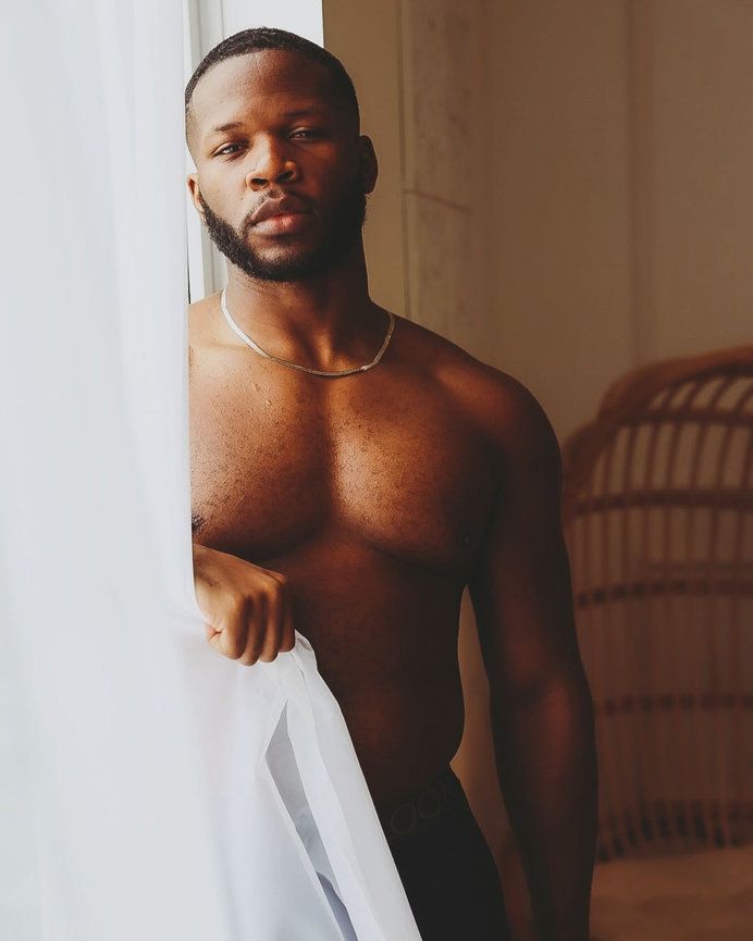 Artistic portrait of a fit model posing shirtless against white curtains in natural lighting.