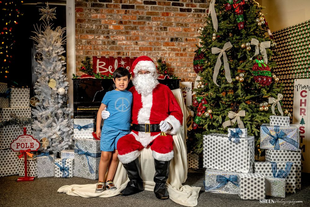 Santa Claus sitting with visitor by Christmas tree surrounded by wrapped gifts.