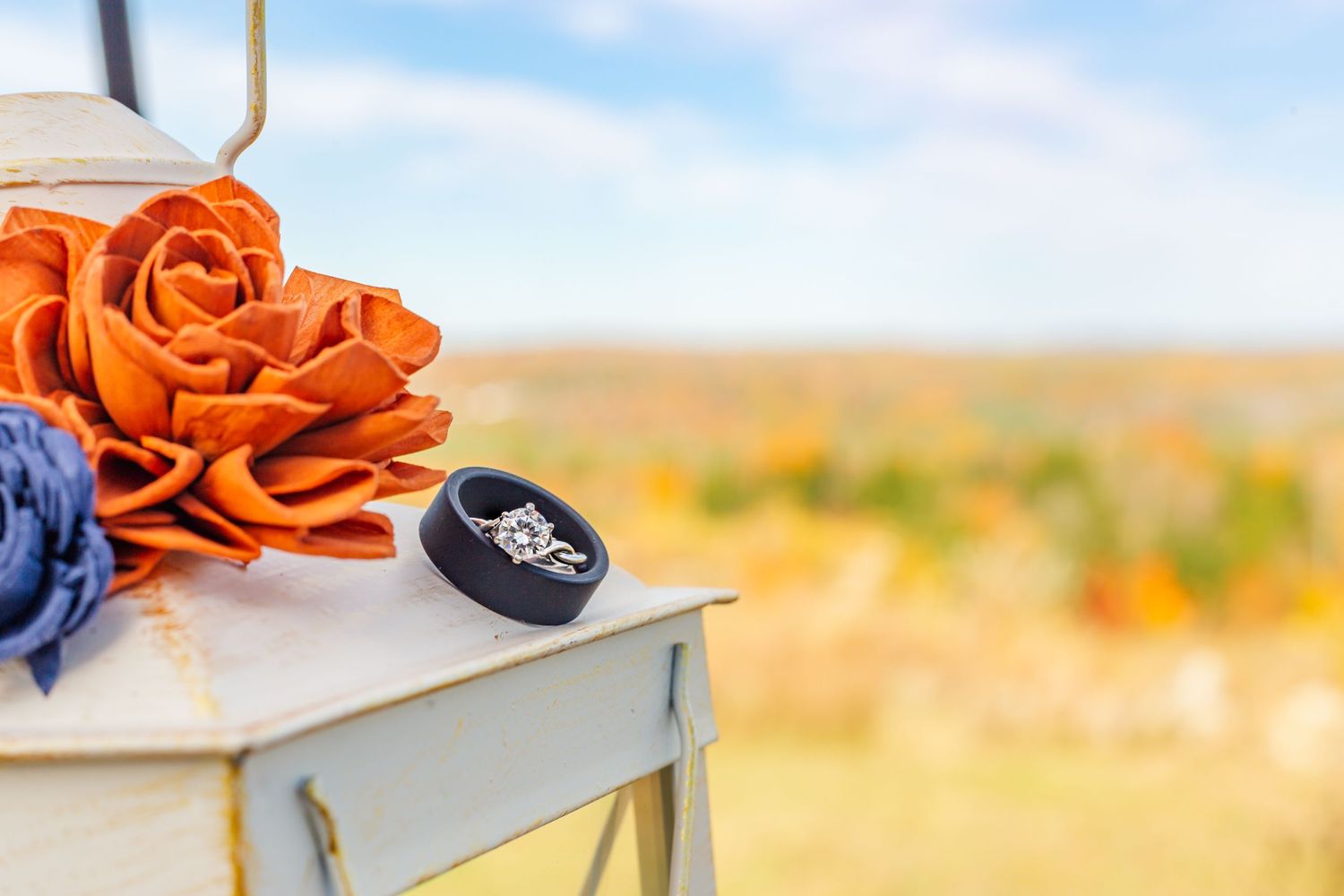 Wedding rings displayed on white surface with orange roses and blue flowers against blurred outdoor backdrop.