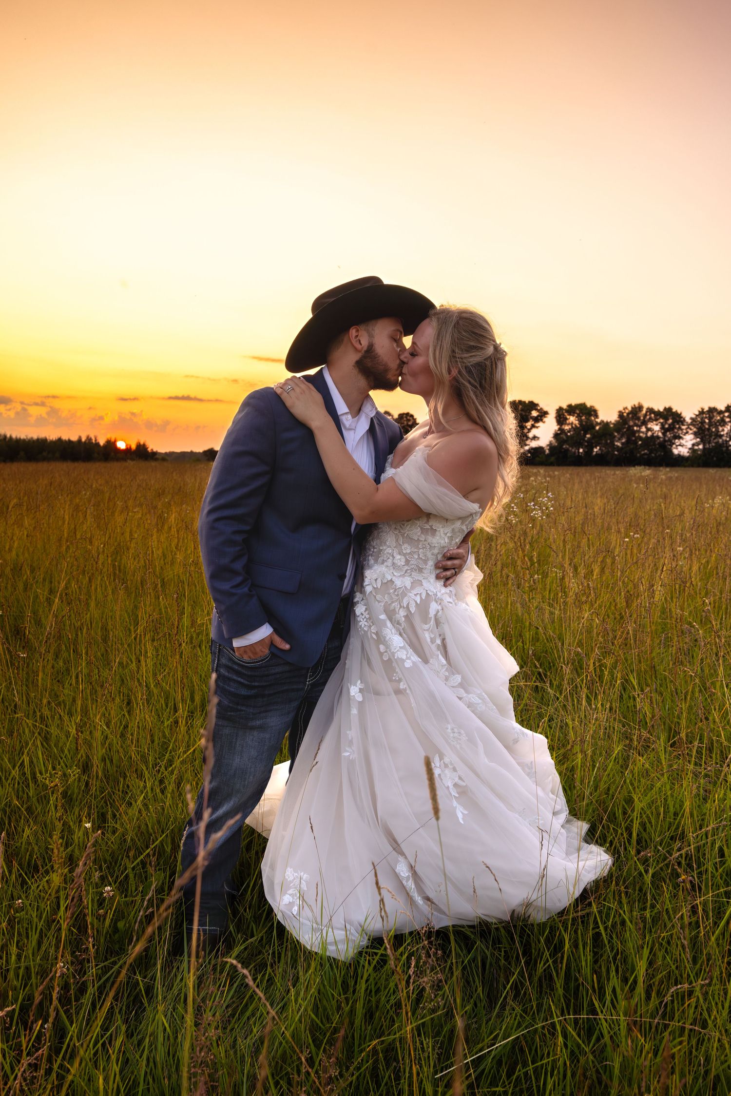 A romantic wedding couple shares an intimate kiss in a golden field at sunset, surrounded by tall grass and warm light.