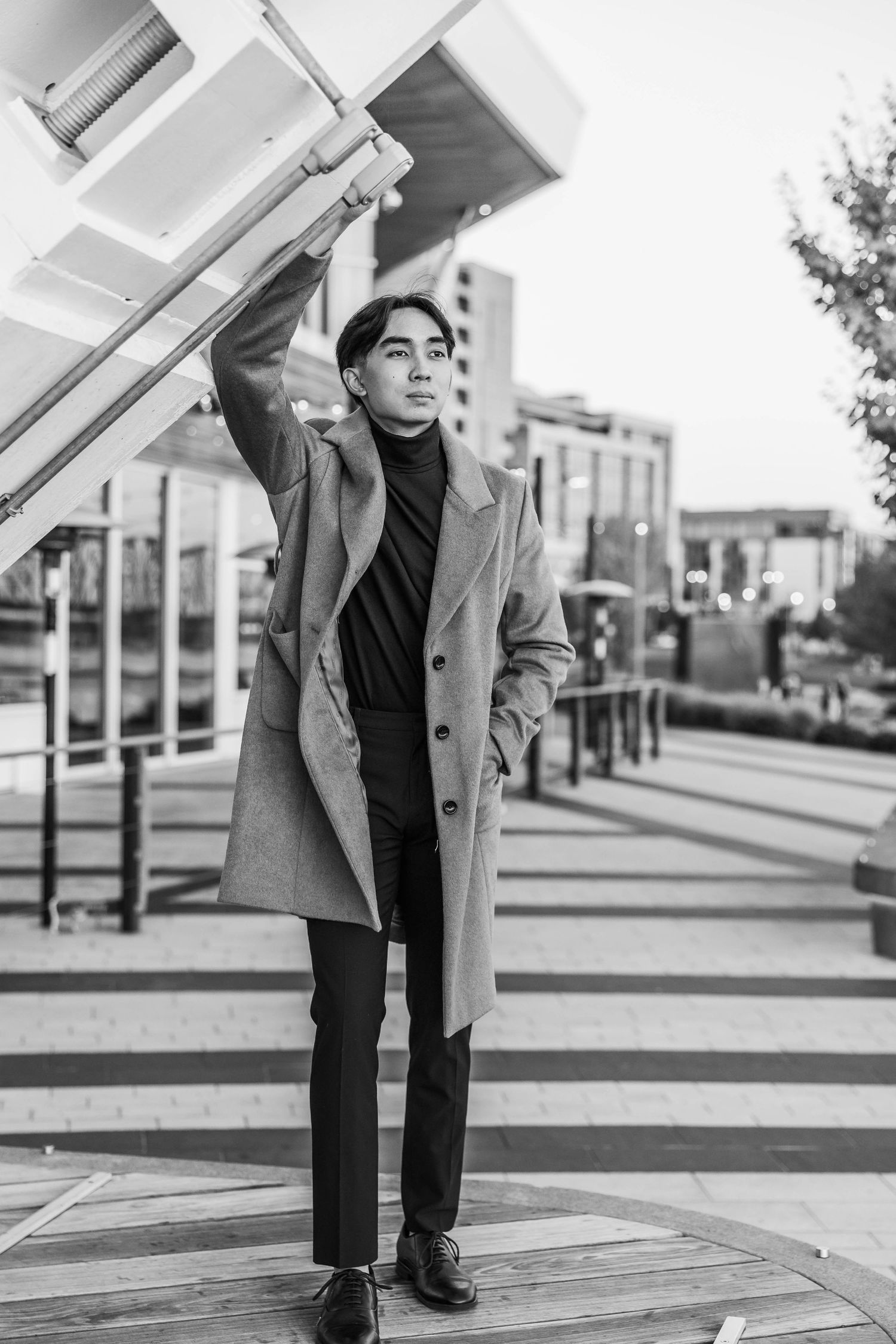 Young Asian man in camel coat reaching up to hold structure in black and white urban portrait.
