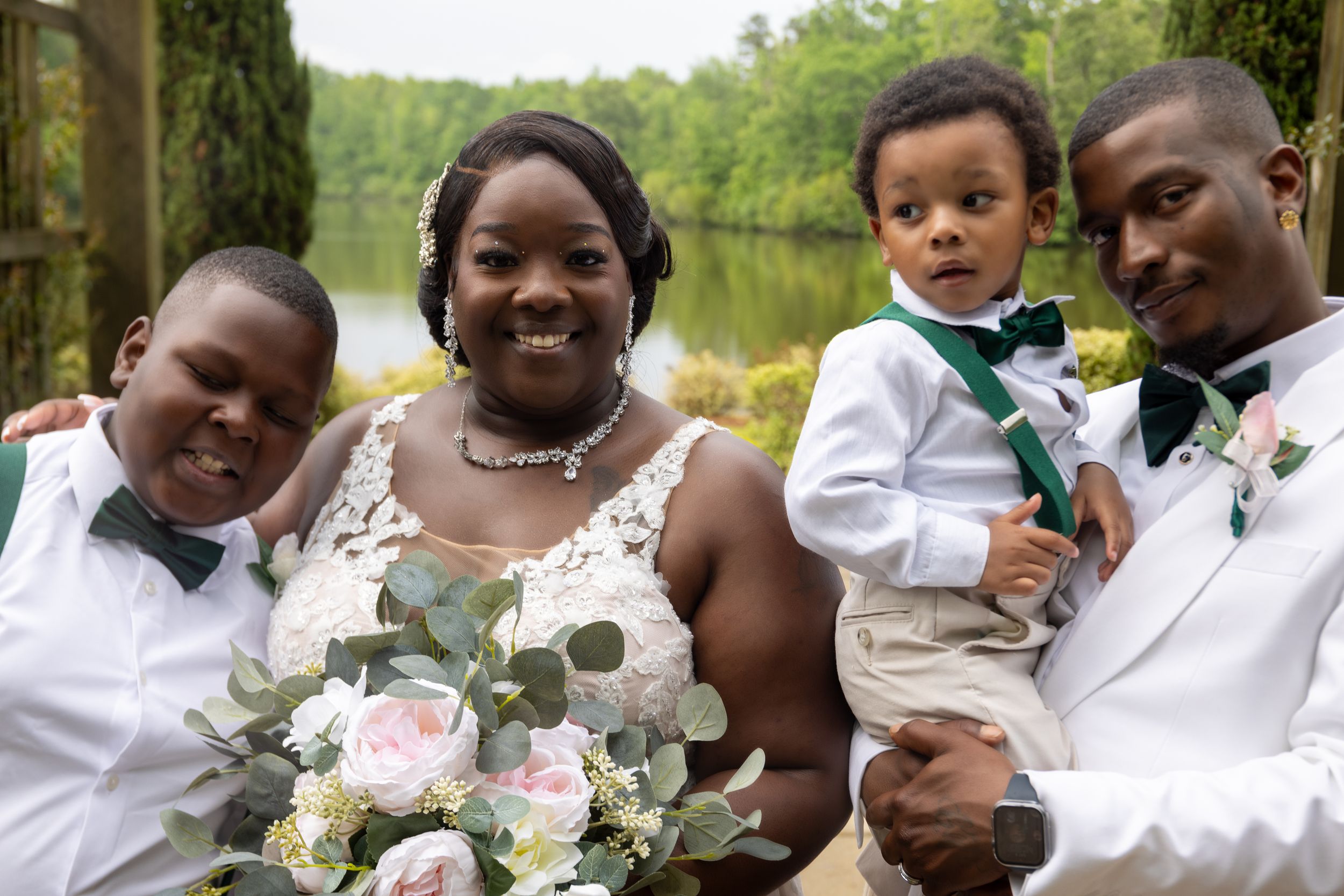 Wedding party in white attire holding floral bouquet by lakeside.