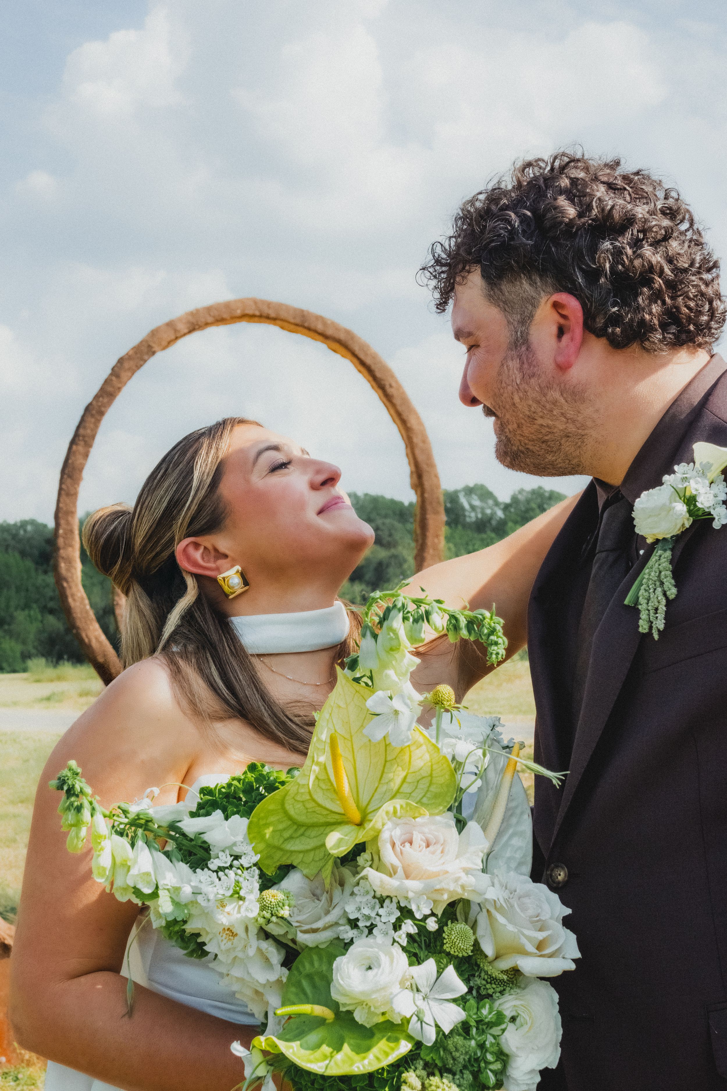 Bride and groom gazing at each other outdoors, bride holding white floral bouquet with green accents near circular wicker arch.