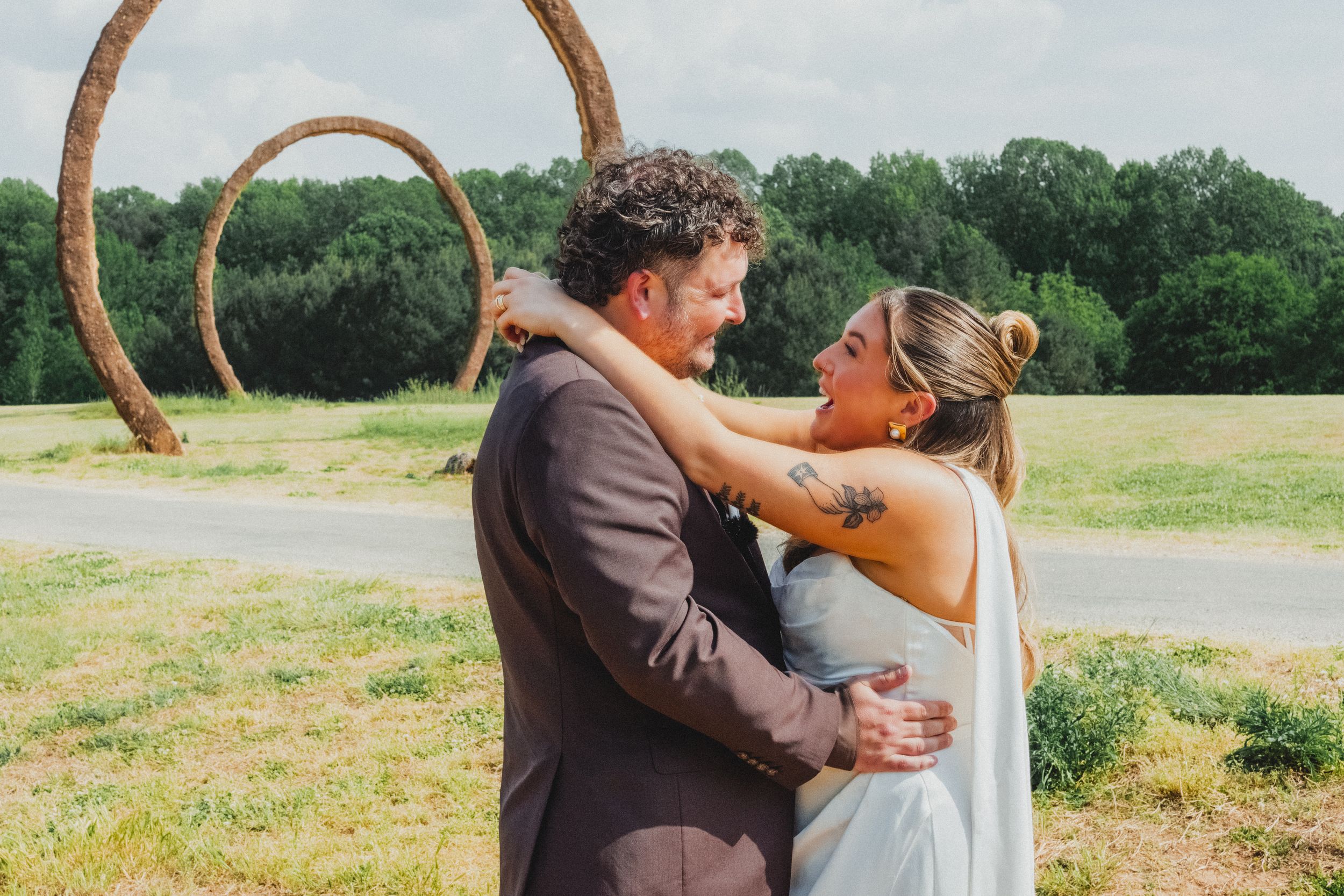 Bride and groom embracing outdoors at wedding venue with rustic circular arch and green trees in background.