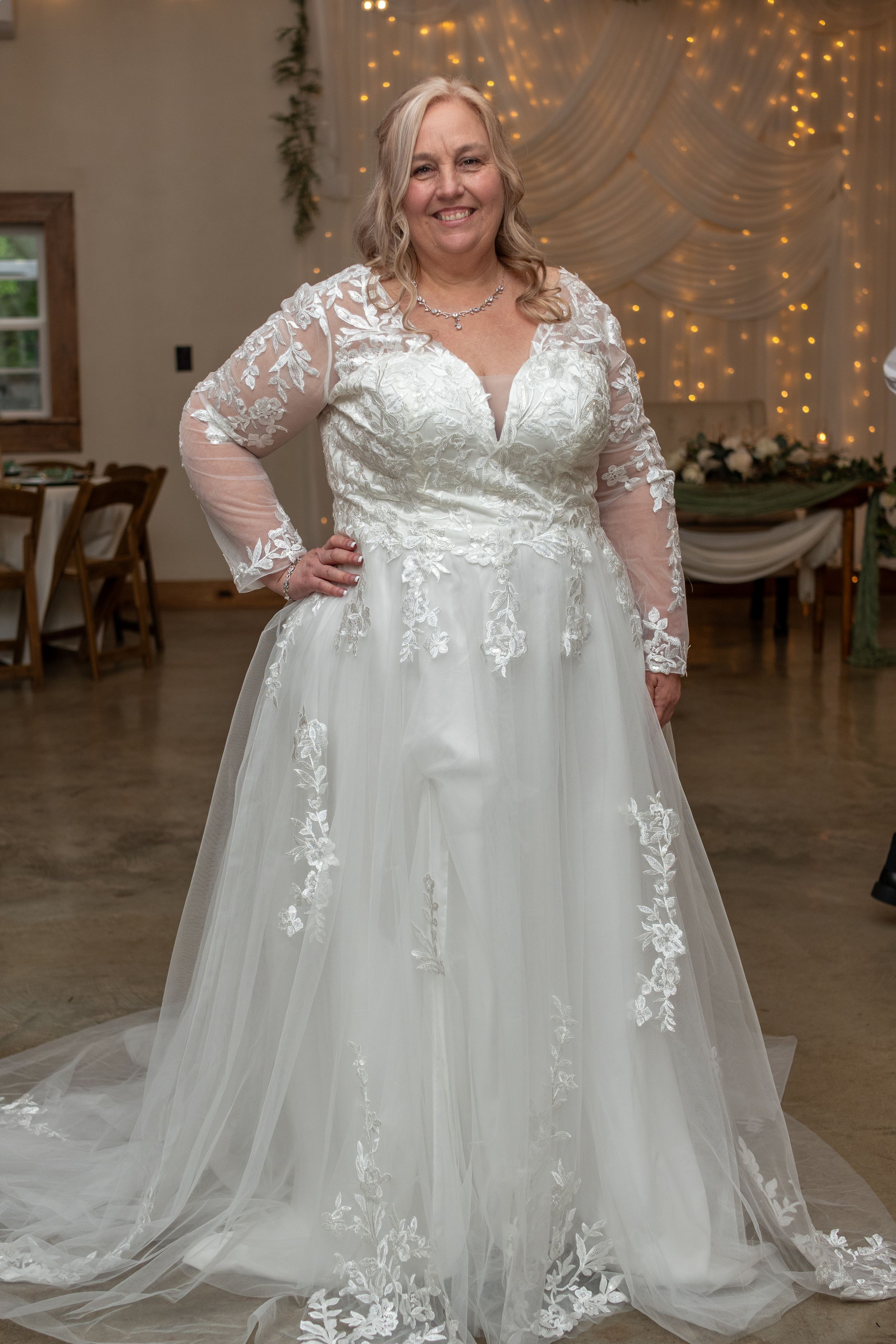 Smiling bride in a long-sleeve lace and tulle A-line wedding gown with floral appliques, posing in an elegant reception venue.