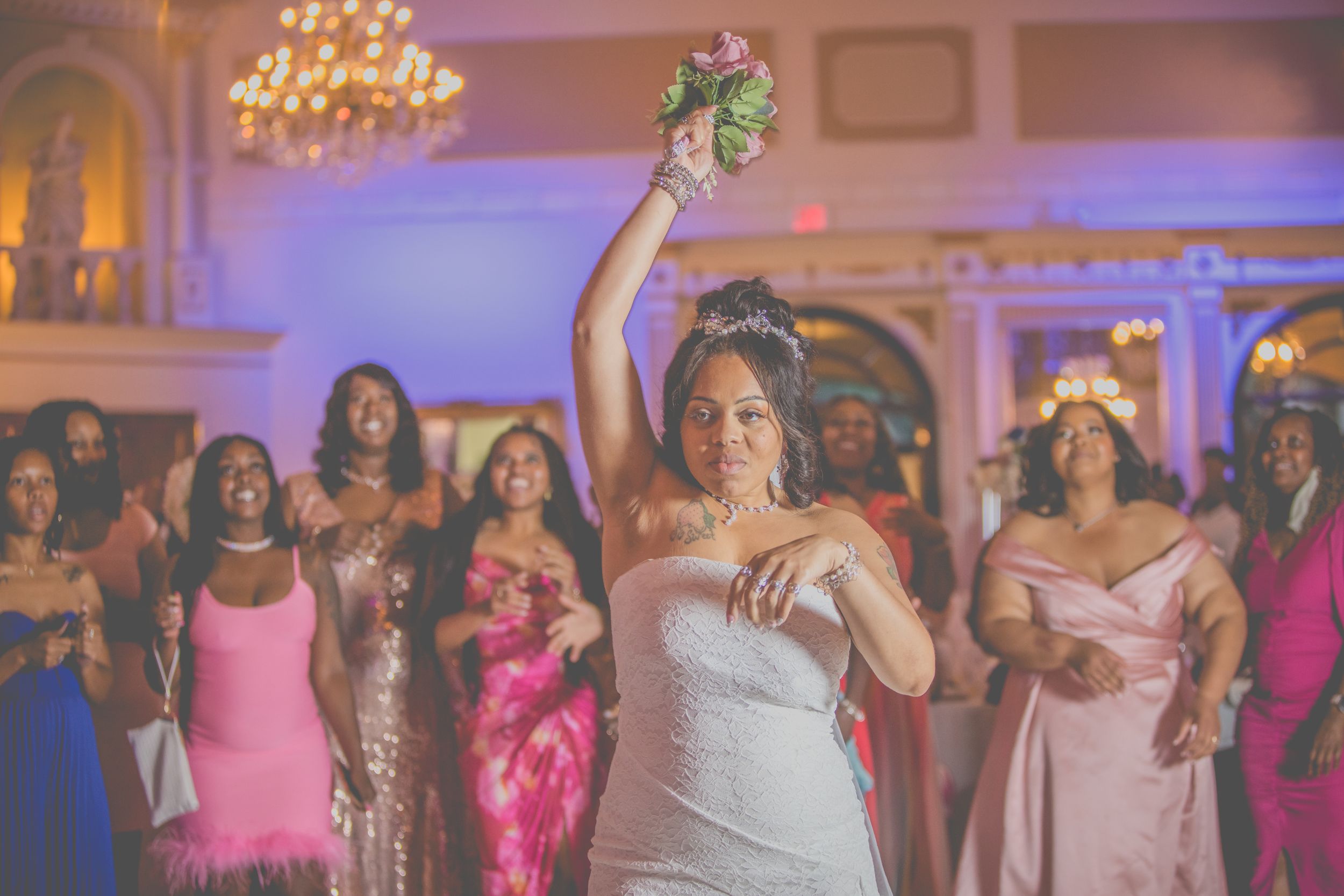 Wedding celebration scene with someone in white dress tossing bouquet while guests in pink dresses watch in elegant ballroom.
