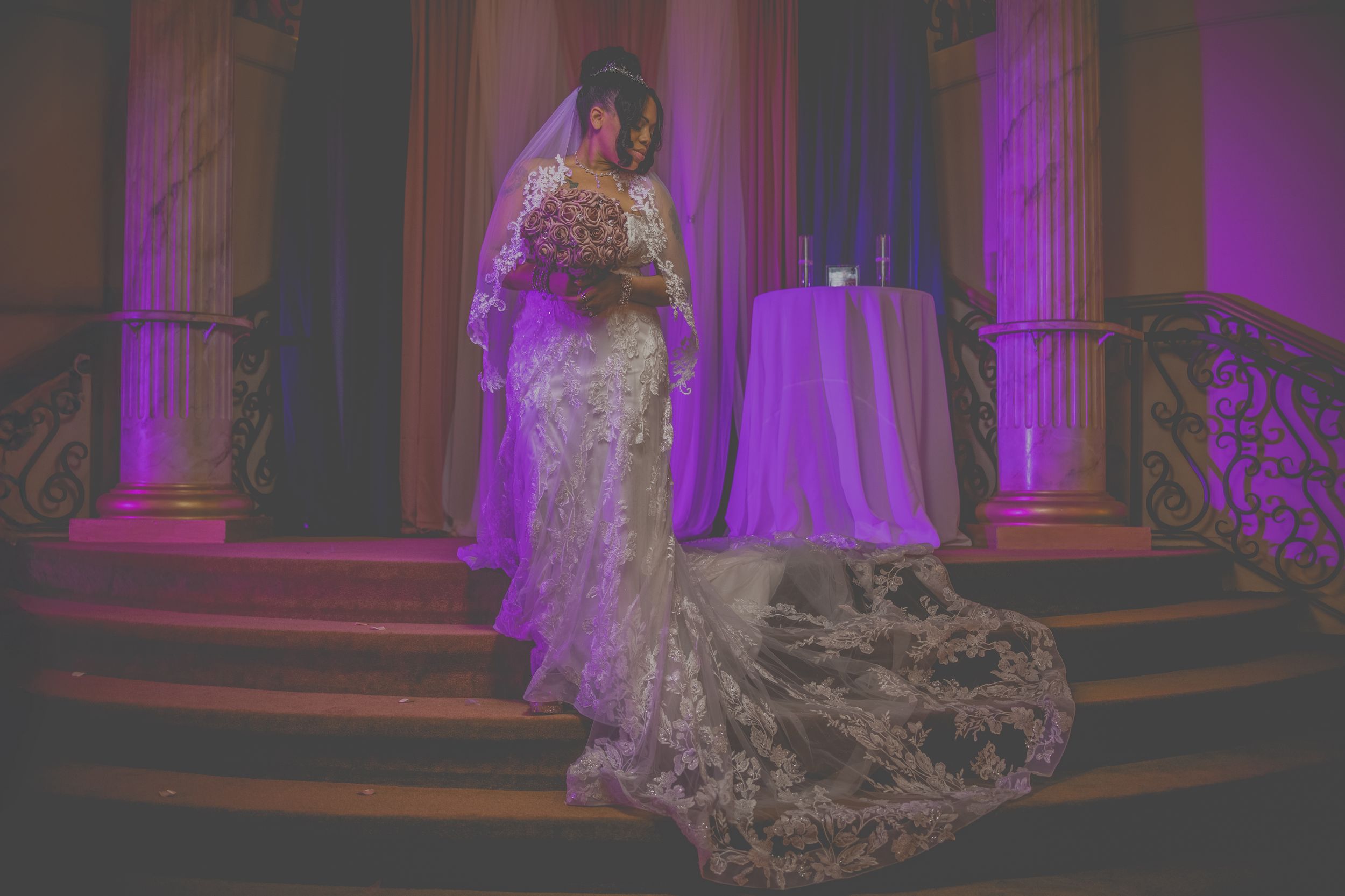 A bride in a white lace wedding gown poses elegantly on a grand staircase illuminated by purple lighting.