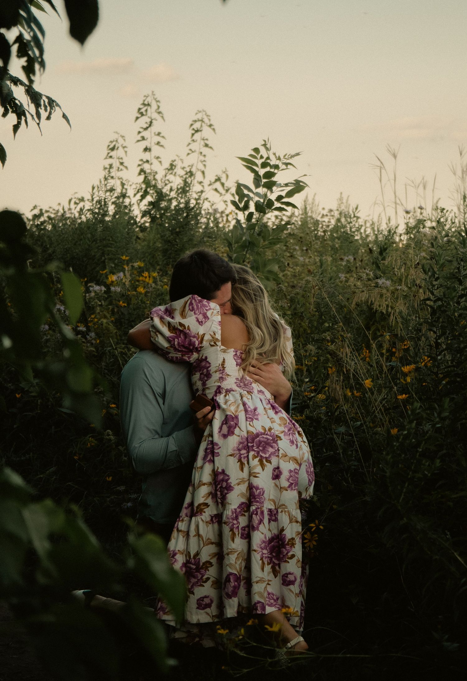 Romantic couple embraces in a wildflower field at sunset, with person wearing floral dress.