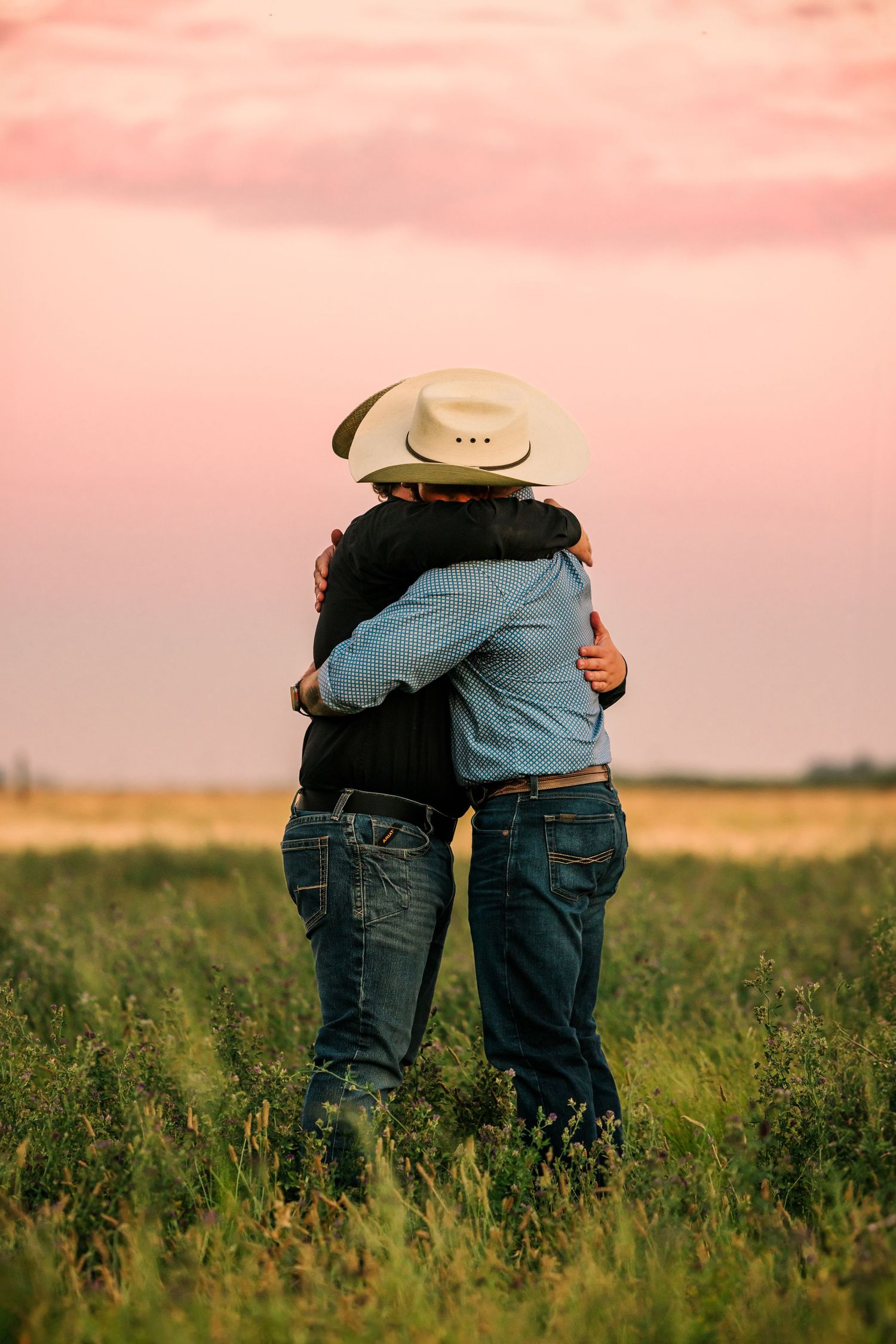Two people embrace in grassy field during sunset wearing western attire.