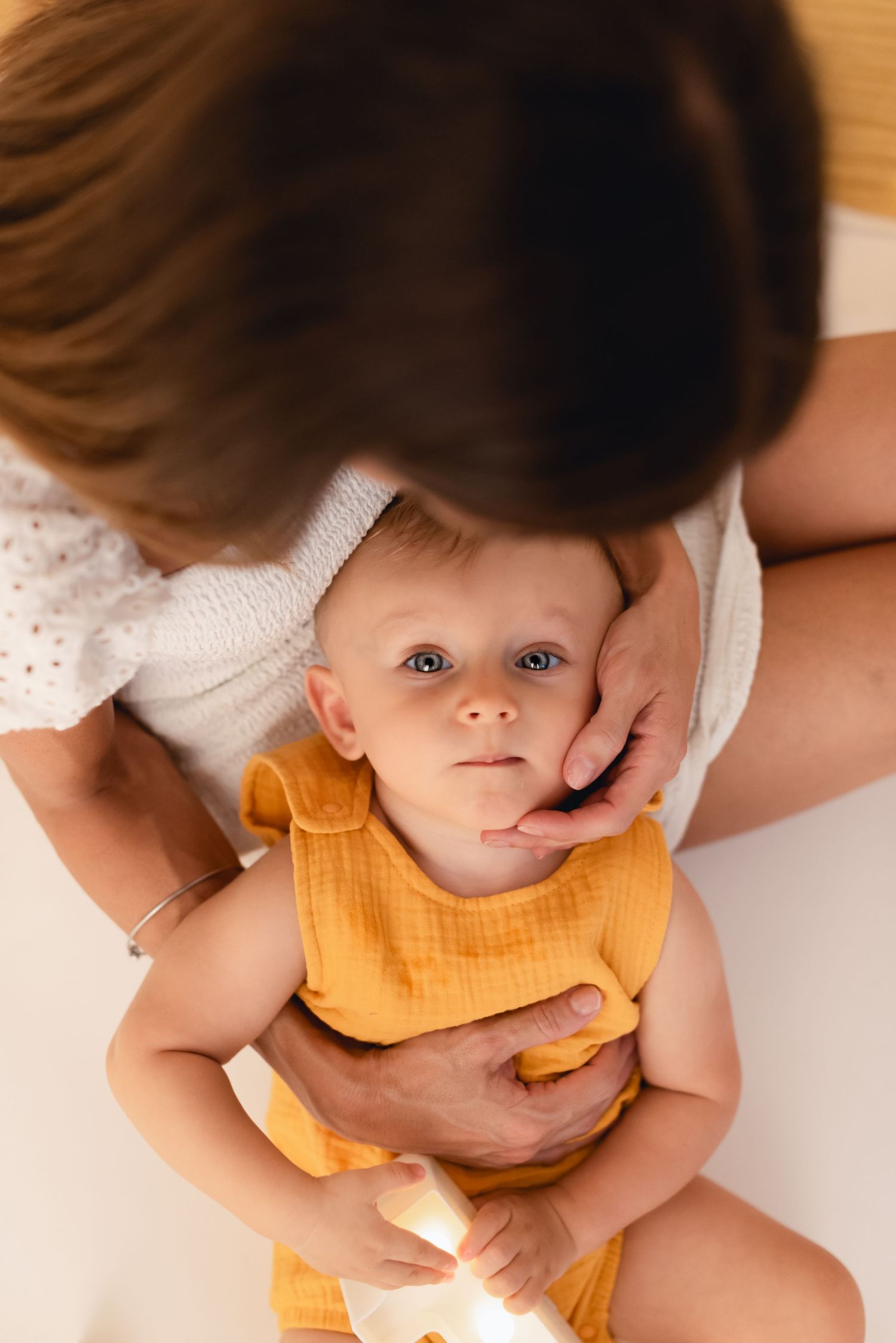 Mother gently cradling a blue-eyed baby in an orange outfit, viewed from above in soft natural light.