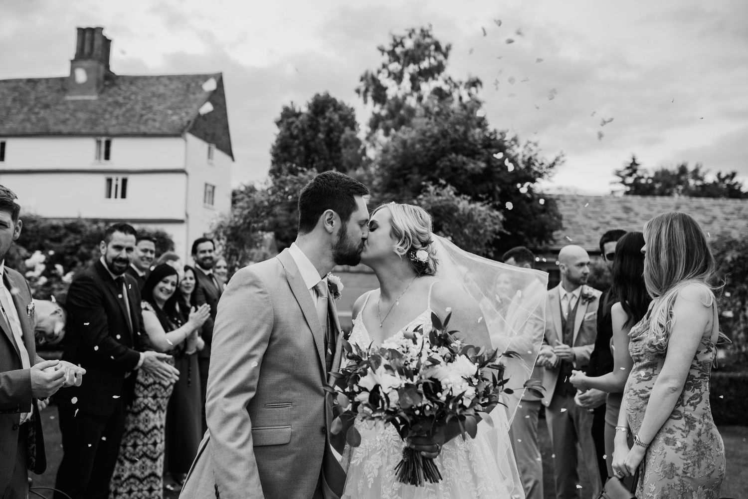 Black and white photo of couple sharing intimate moment at garden wedding with guests in background. Houchins wedding venue confetti throw