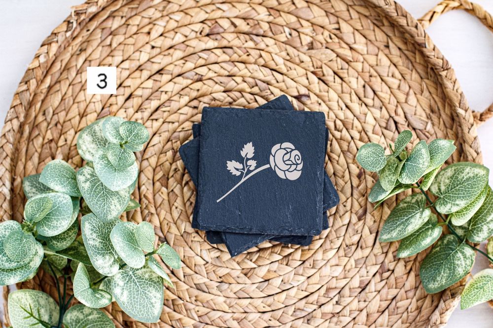 Navy blue coaster with white rose design placed on circular woven mat with eucalyptus accents.
