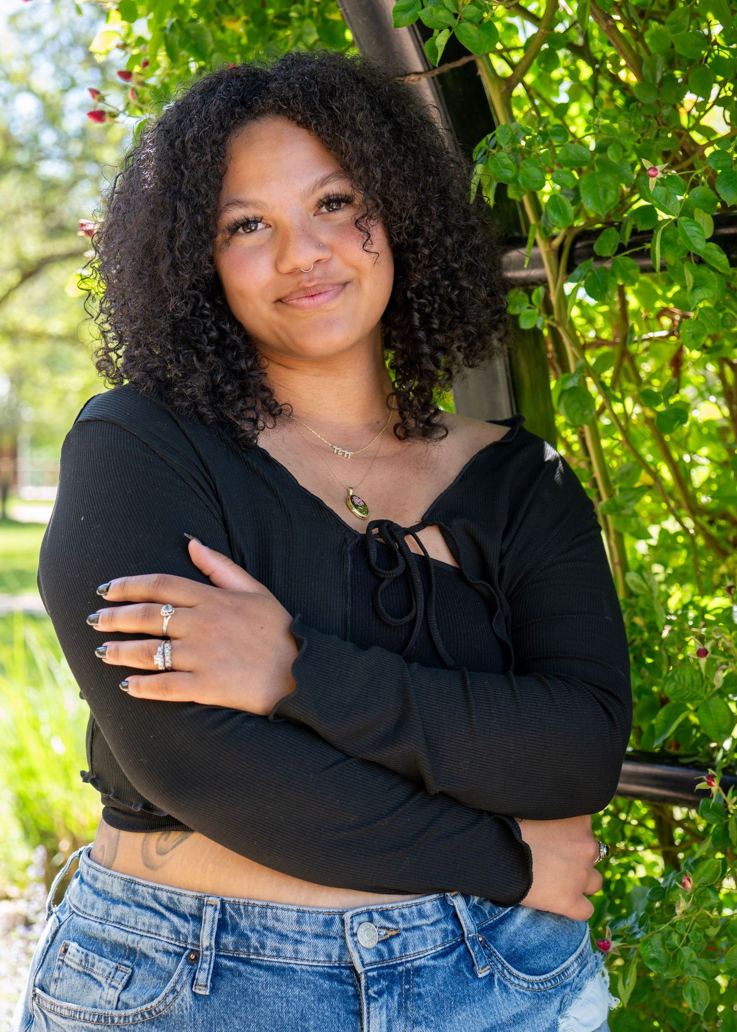 Young woman with curly hair in black top and jeans poses outdoors with arms crossed near green foliage.