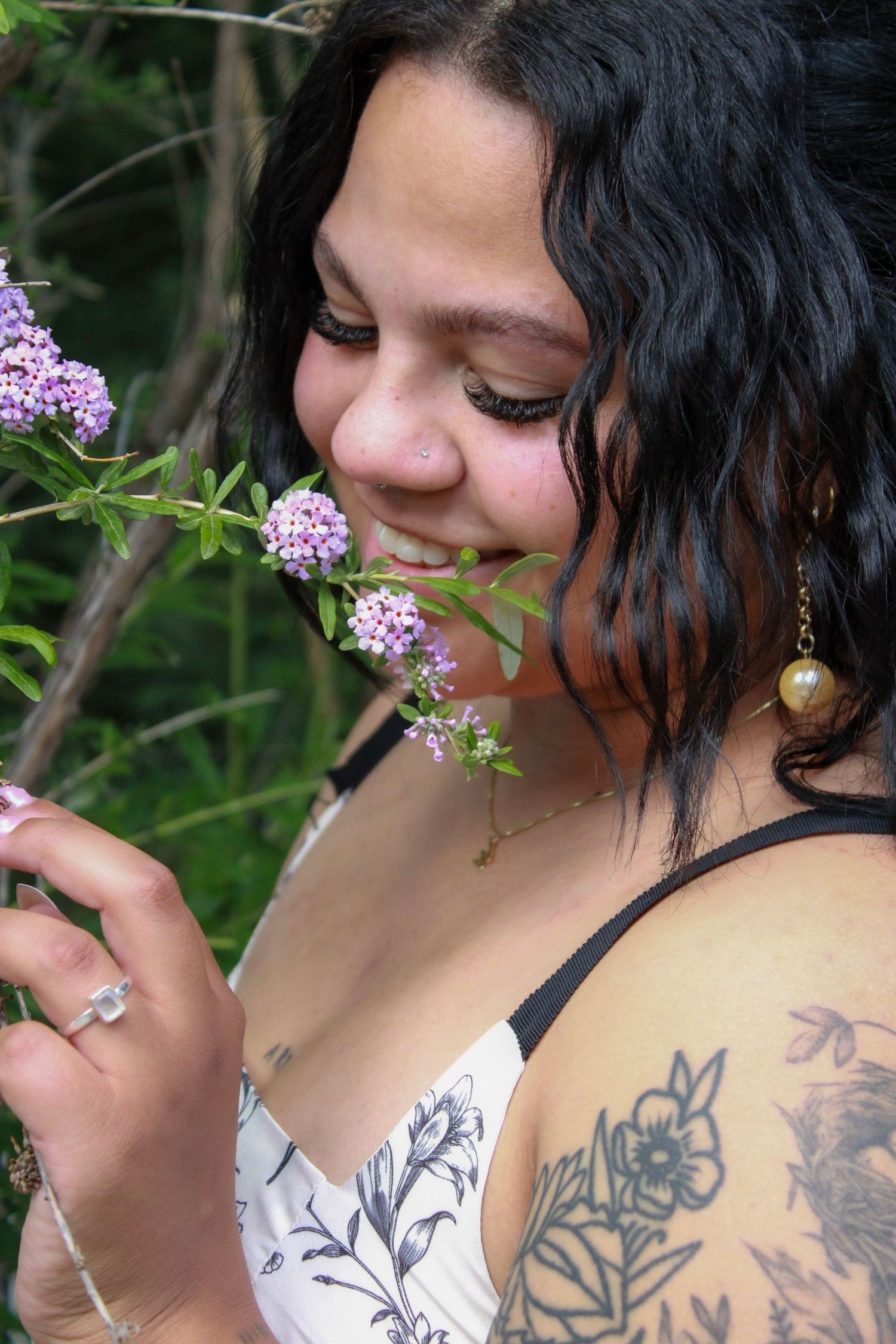 Young woman with black hair and floral tattoo smells purple wildflowers outdoors, wearing a white floral dress.