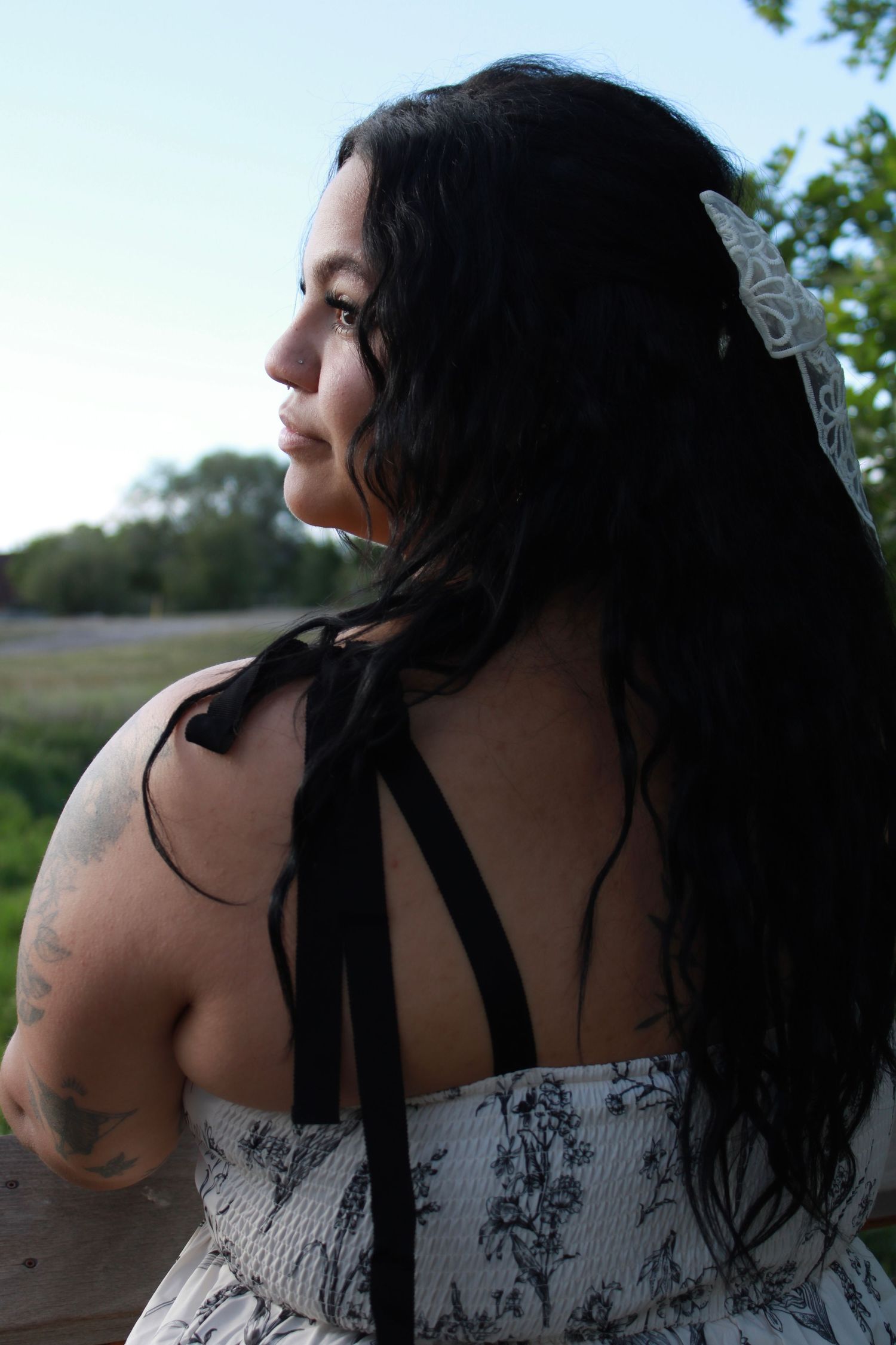 Woman with dark hair and white bow accessory wearing floral halter dress with black ribbon ties, viewed from behind outdoors.