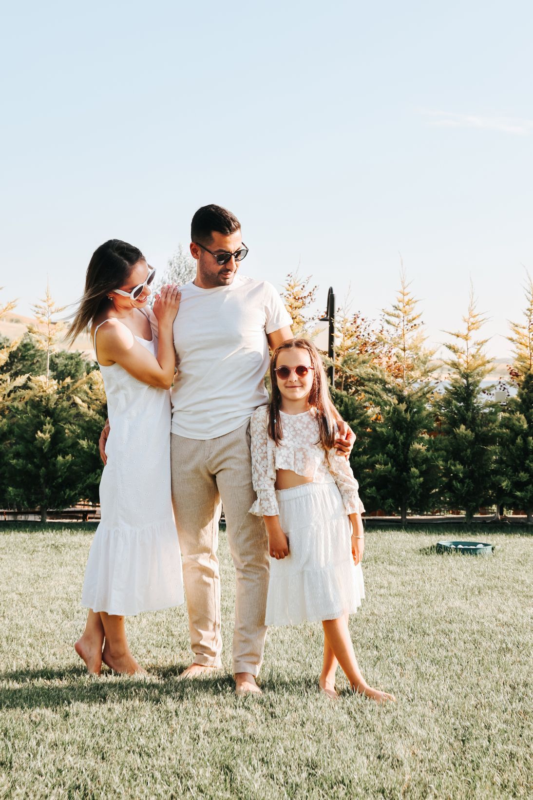 Family in coordinated white outfits standing together in a sunny garden with evergreen trees.