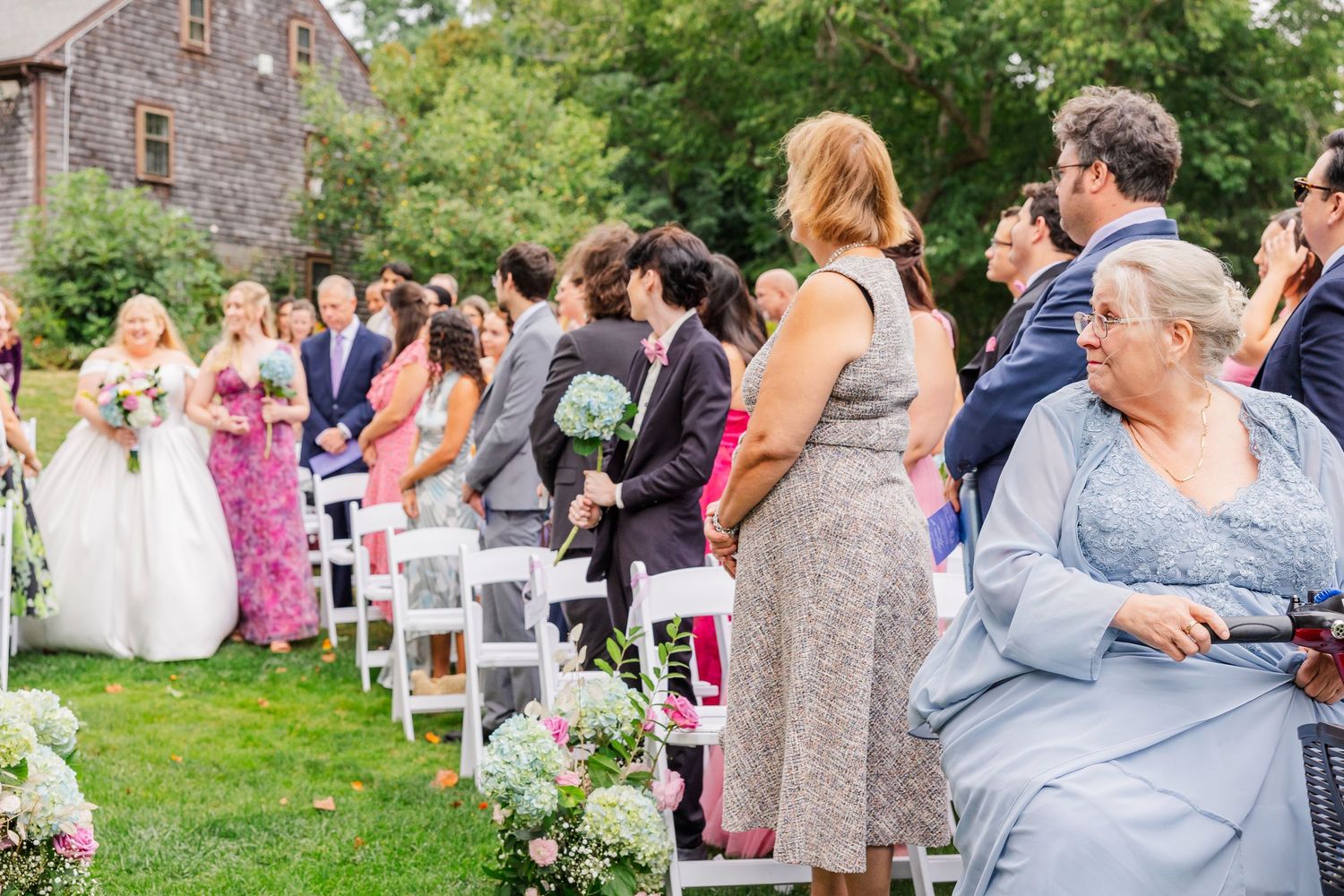 Wedding guests seated in white chairs during an outdoor garden ceremony at a stone building venue.