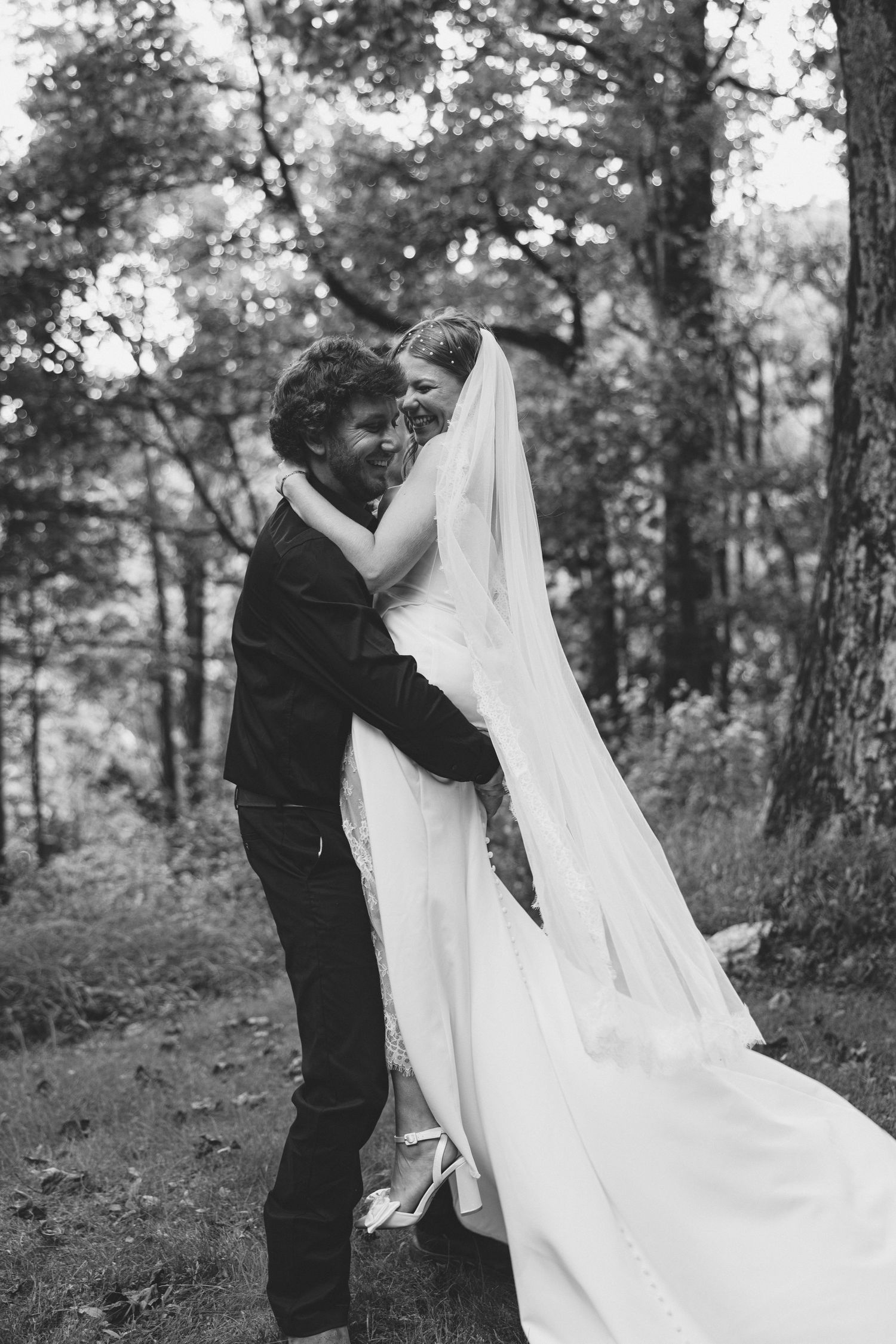A black and white wedding photo showing a couple embracing outdoors in a wooded setting with a flowing white veil. Wedding Photographer