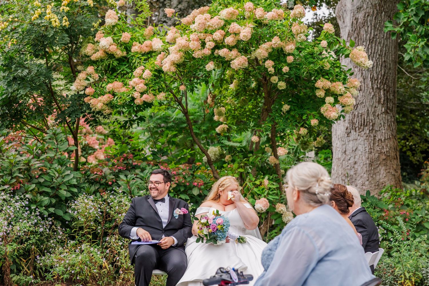 Wedding ceremony takes place in a lush garden setting with hydrangea blooms and tall trees creating a romantic atmosphere.