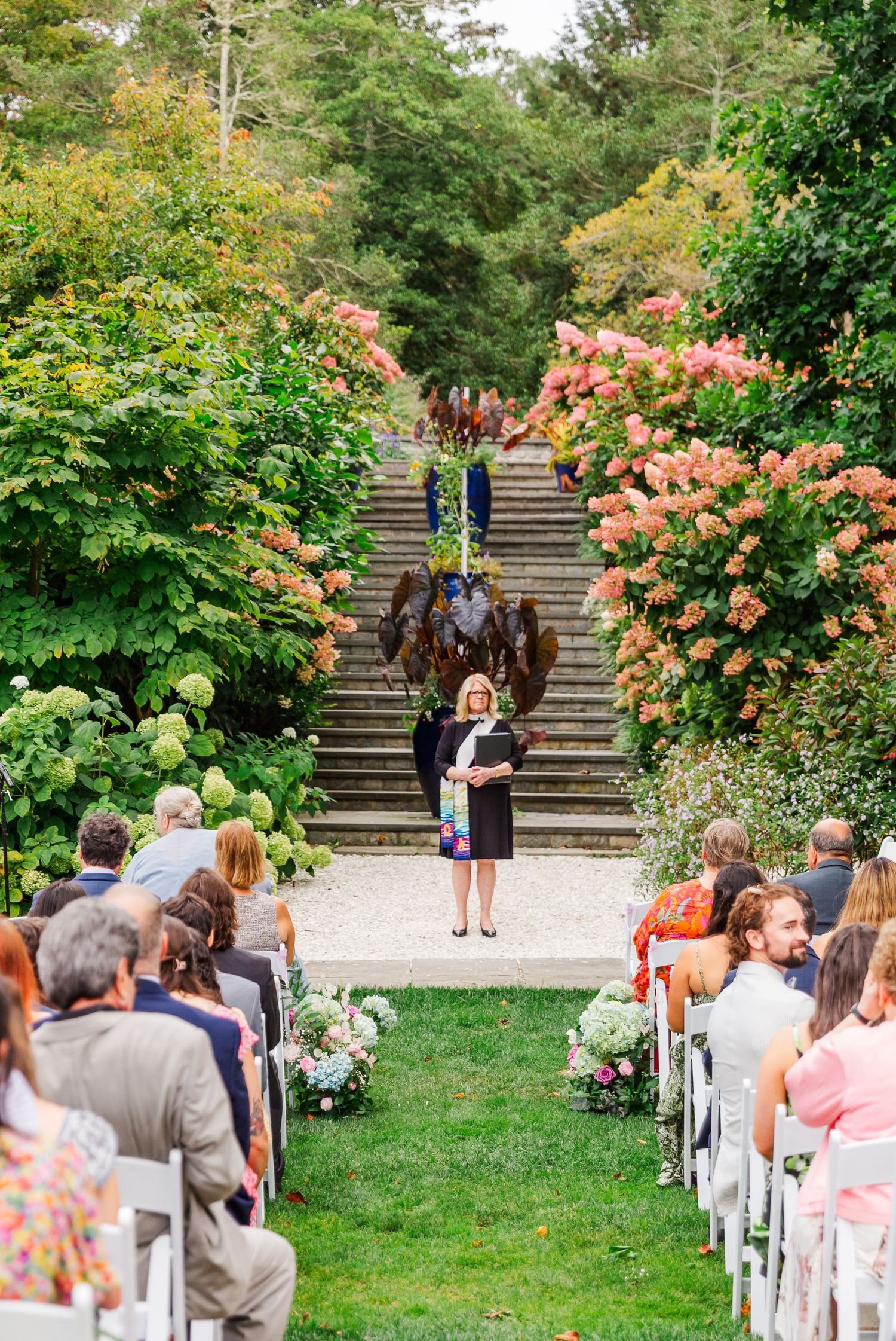 Wedding guests seated in white chairs along garden path with blooming pink roses and stone steps in outdoor setting.