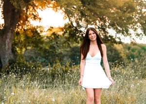Young woman in white dress standing in sunlit meadow with wildflowers and trees in golden hour light.