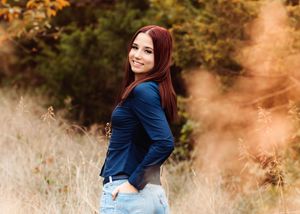 A woman in a blue blazer and light jeans poses outdoors in a golden autumn field during sunset.