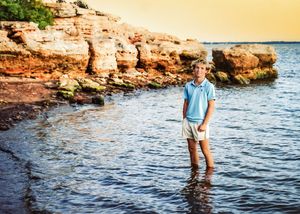 Someone wades in shallow coastal waters near rocky red cliffs during golden hour at sunset.